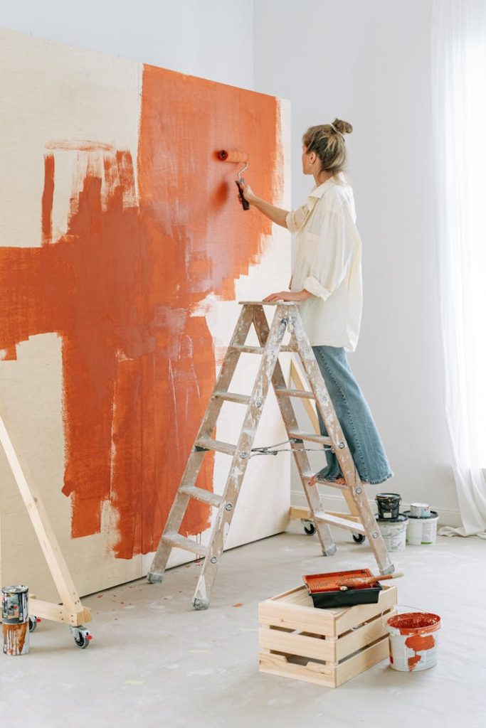 pexels-photo-5799043 A woman stands on a stepladder, painting a room's wall during home renovation.